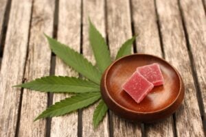 a small round dish containing two cannabis gummies on top of a cannabis leaf over a slat wood table.
