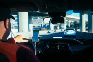 The interior of a car, likely a rideshare vehicle, with a driver and a smartphone mounted on the dashboard, displaying a map or navigation.