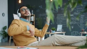 A man in a yellow sweater and khaki pants listening to music through headphones while sitting in an office in front of a huge blackboard.